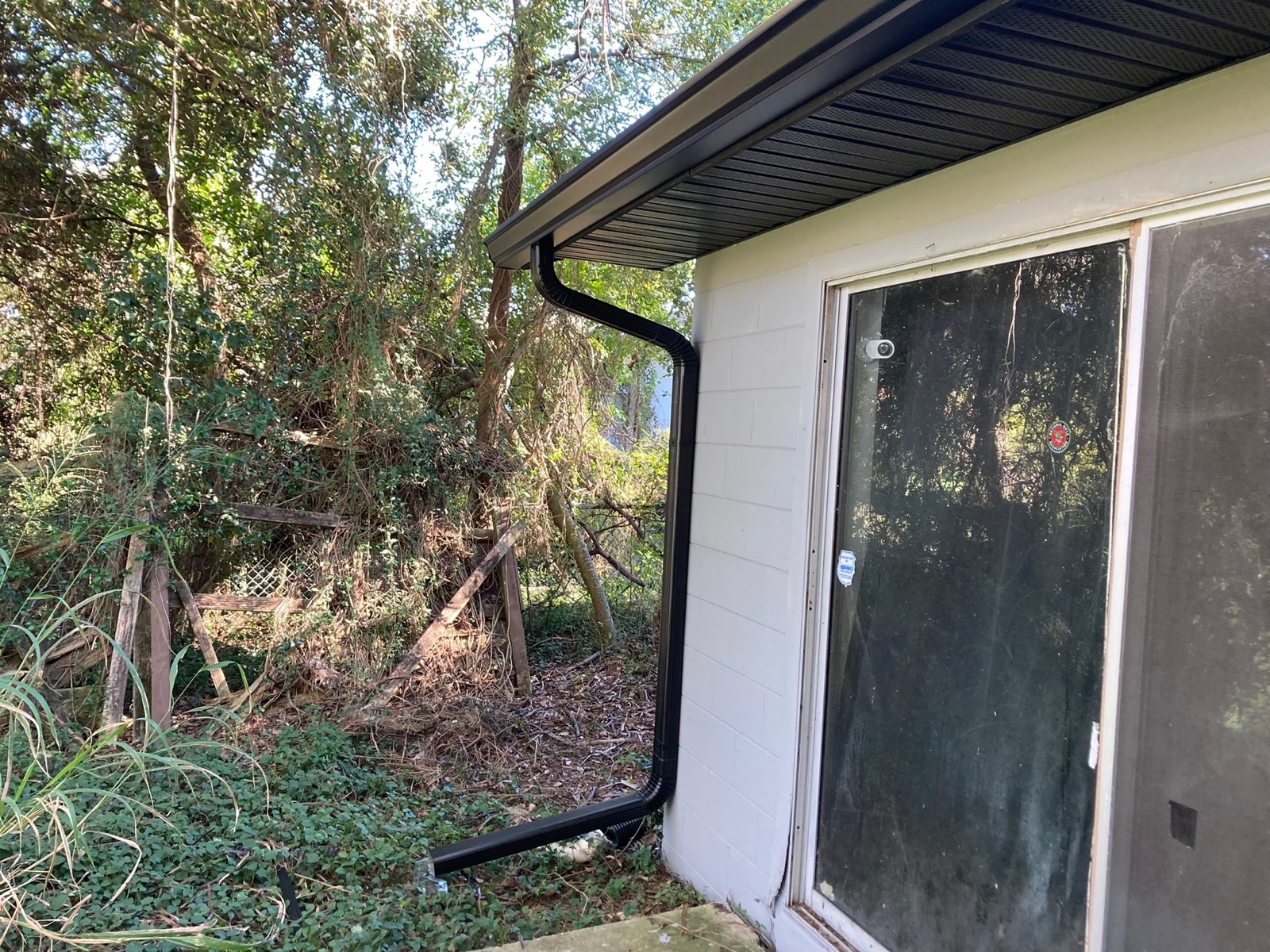 Black downspout retrofit against white siding next to dense foliage